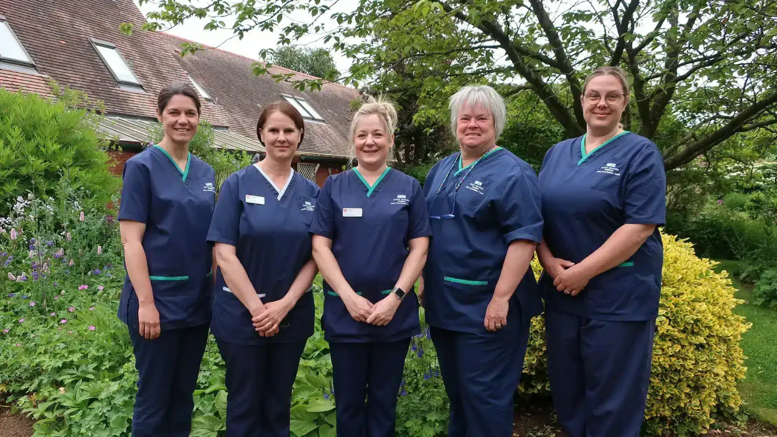 Five palliative care nurses at Katharine House hospice in navy scrubs stand together in the hospice garden, smiling.