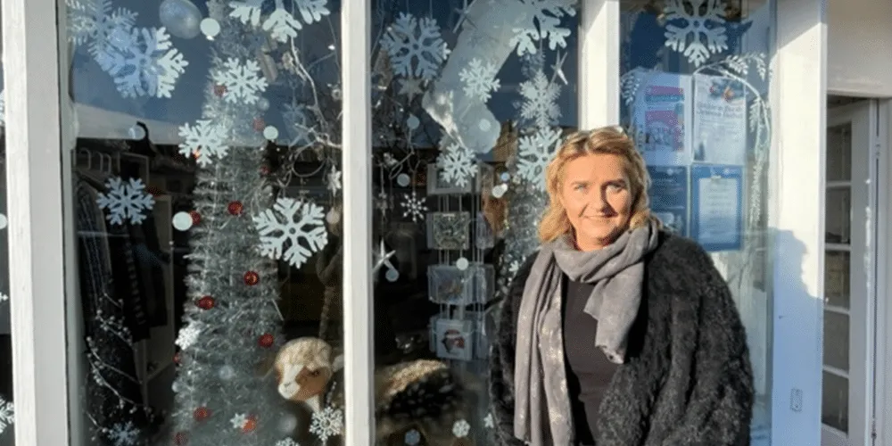Moreton-in-Marsh detail Woman standing in front of a festive window in the Moreton-in-Marsh Katharine House Hospice charity shop with snowflake decorations and a silver tree.