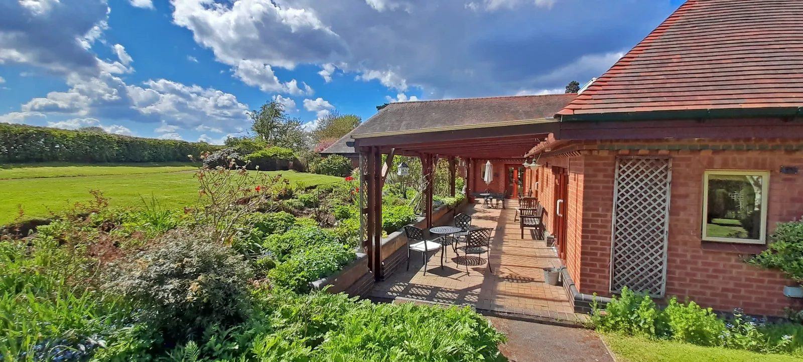 Become a Friend The back of Katharine House Hospice built of brick with a covered patio, set beside the gardens and open green fields under a blue sky.