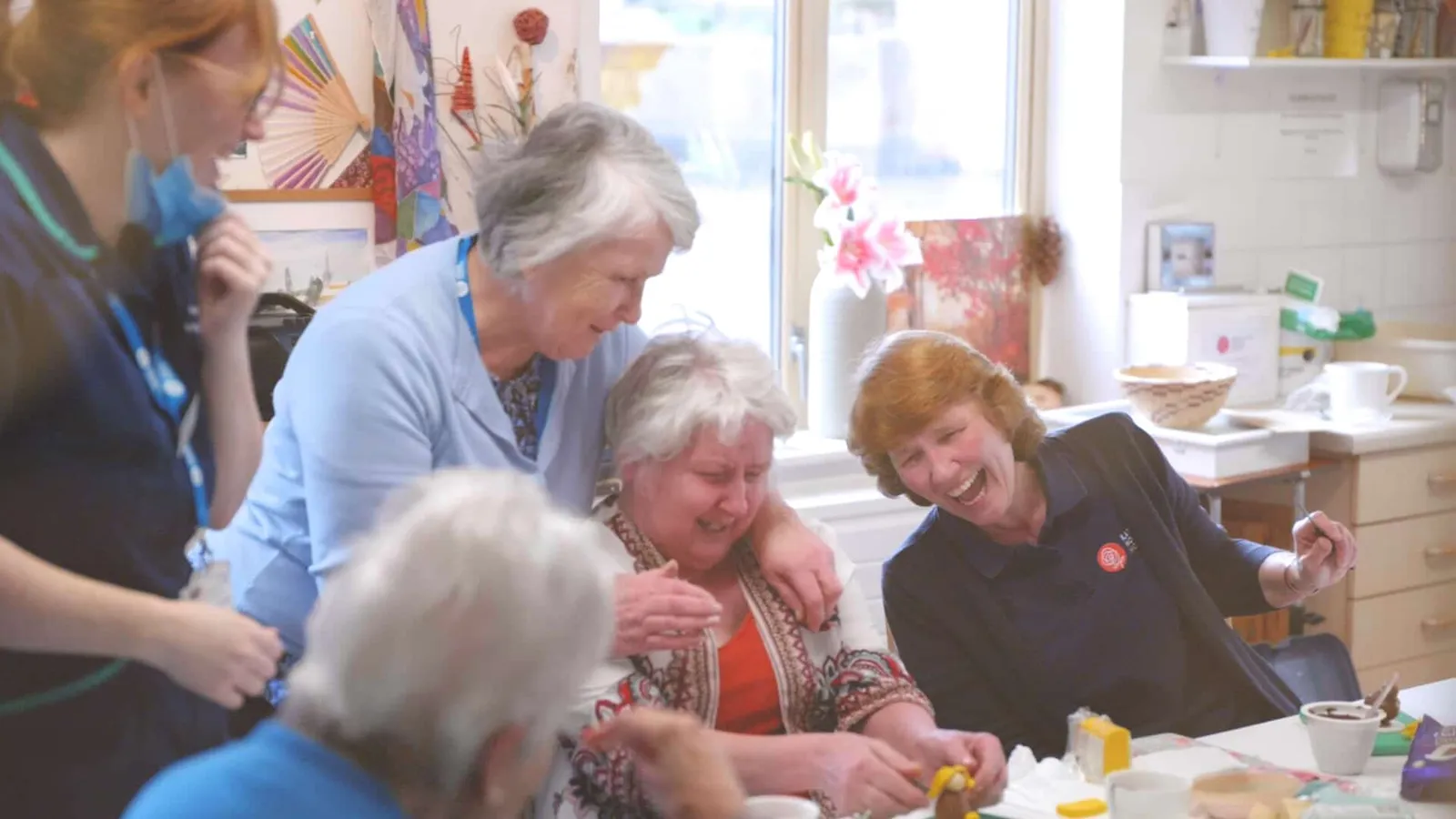 People laughing and enjoying a social gathering around a table in the Living Well lounge at Katharine House Hospice.
