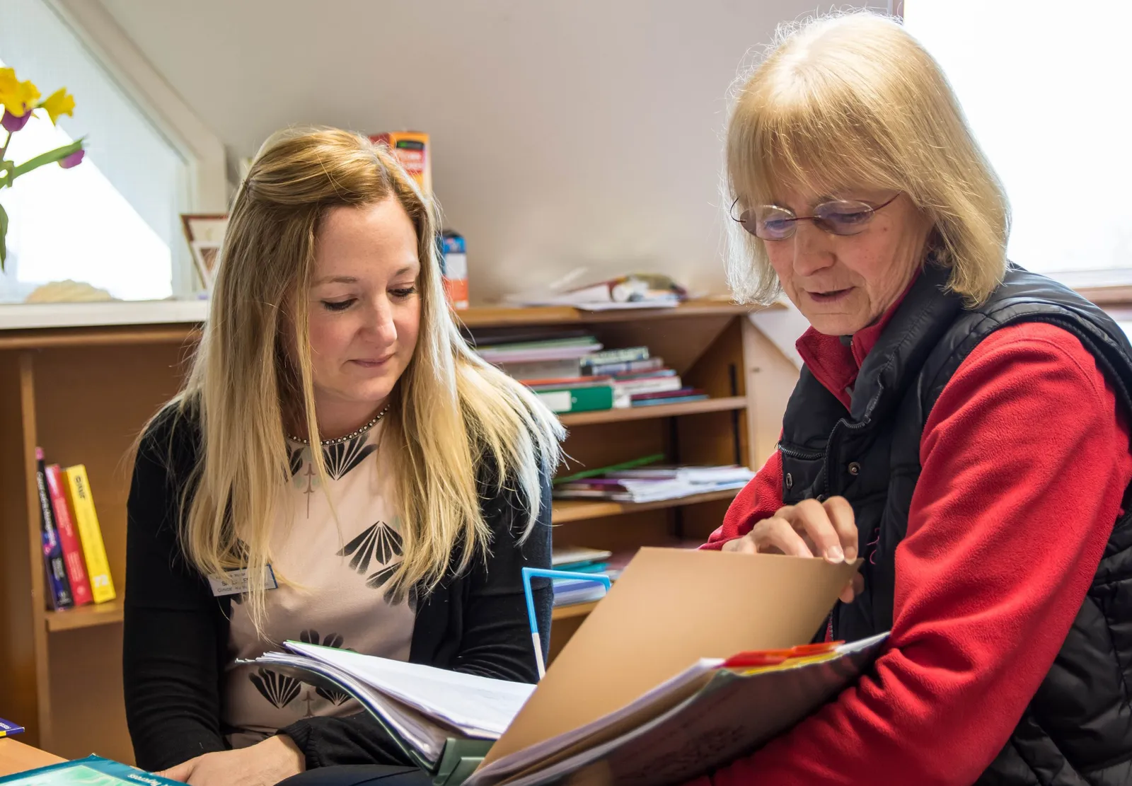 Two support workers in an office at Katharine House Hospice with shelves of books behind them viewing a file.